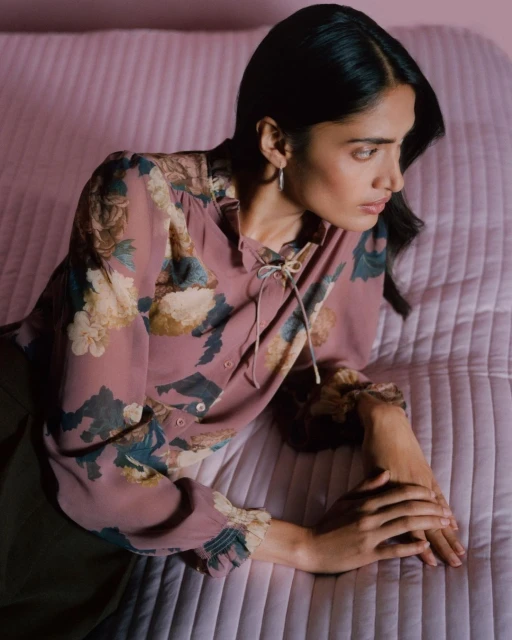 Portrait of a woman with dark hair wearing a floral blouse, resting her hands on a textured pink surface in an indoor setting.