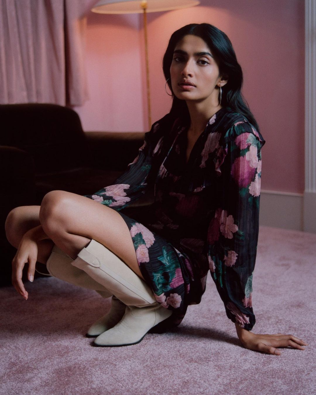 Dark-haired adult female in floral-print dress and cream-colored boots poses in a crouching position on a pink carpet indoors.