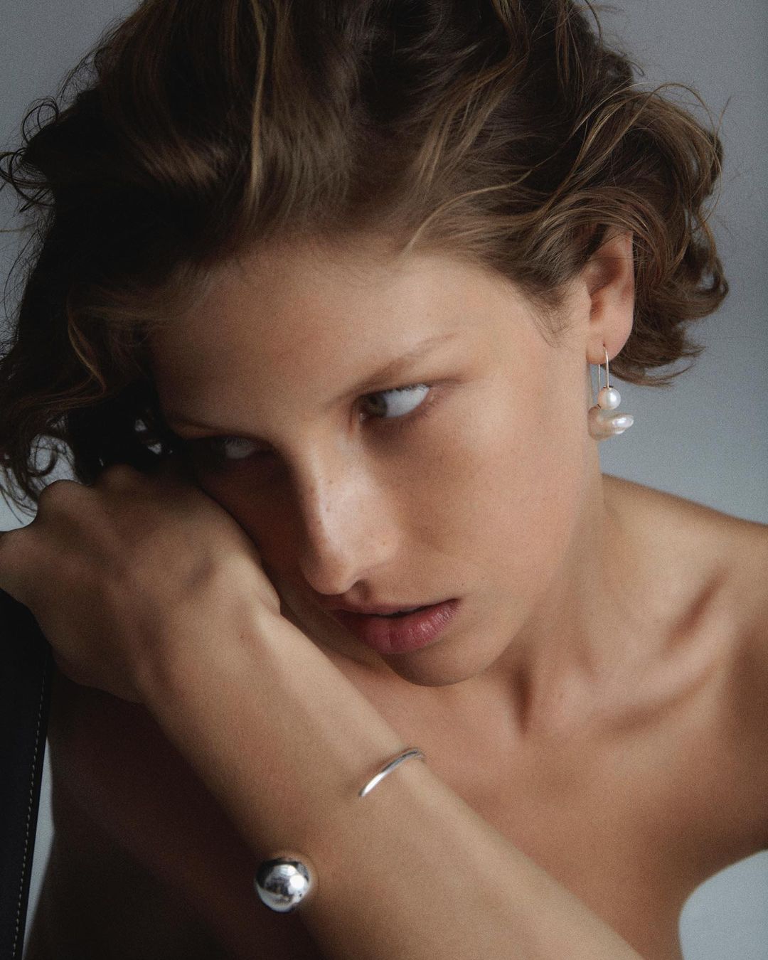 Close-up portrait of a young adult woman with short brown hair wearing pearl earrings and silver bracelets against a neutral background.