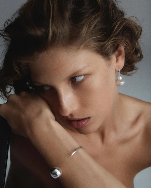 Close-up portrait of a young adult woman with short brown hair wearing pearl earrings and silver bracelets against a neutral background.