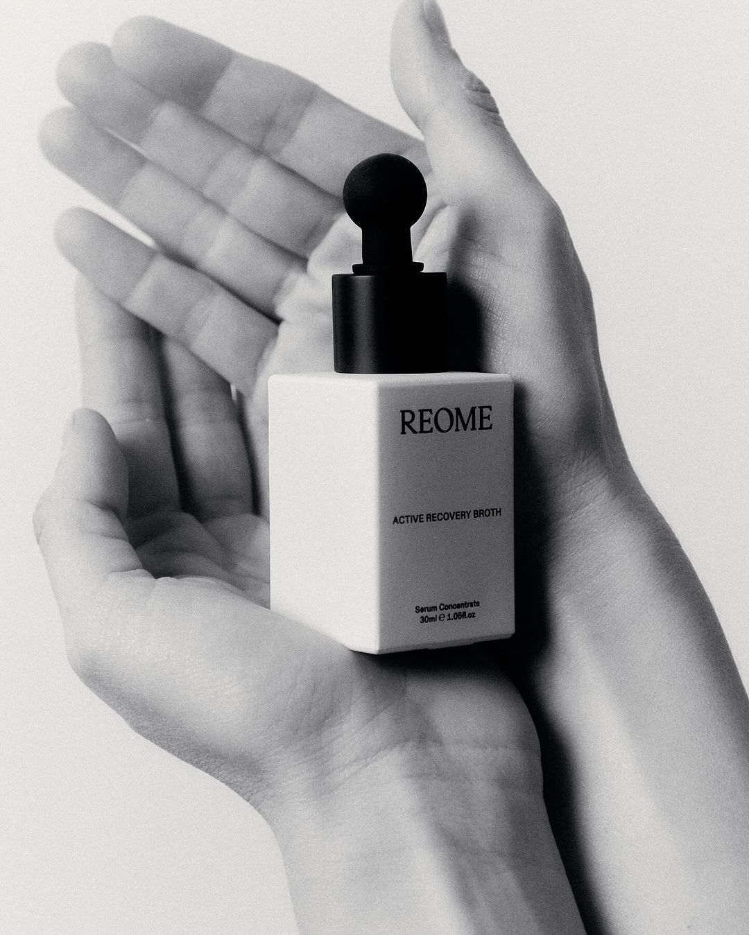 Monochrome close-up of skincare product held in two hands, showcasing product's cube bottle and dropper against a light backdrop.