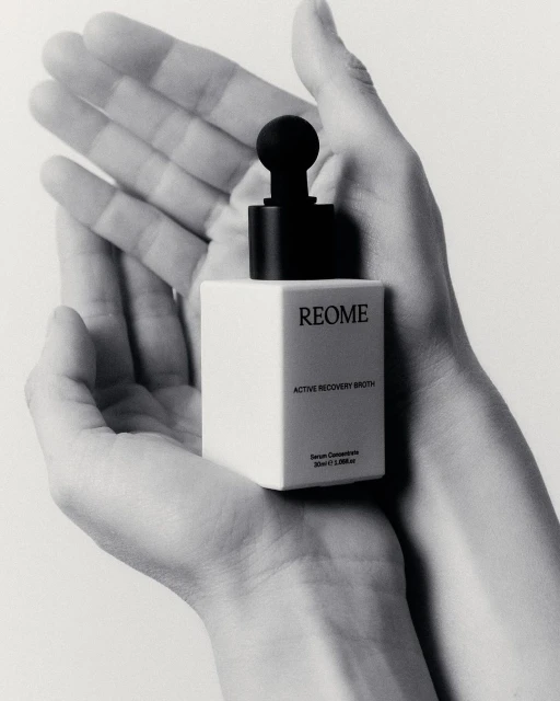 Monochrome close-up of skincare product held in two hands, showcasing product's cube bottle and dropper against a light backdrop.