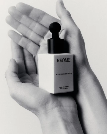 Monochrome close-up of skincare product held in two hands, showcasing product's cube bottle and dropper against a light backdrop.