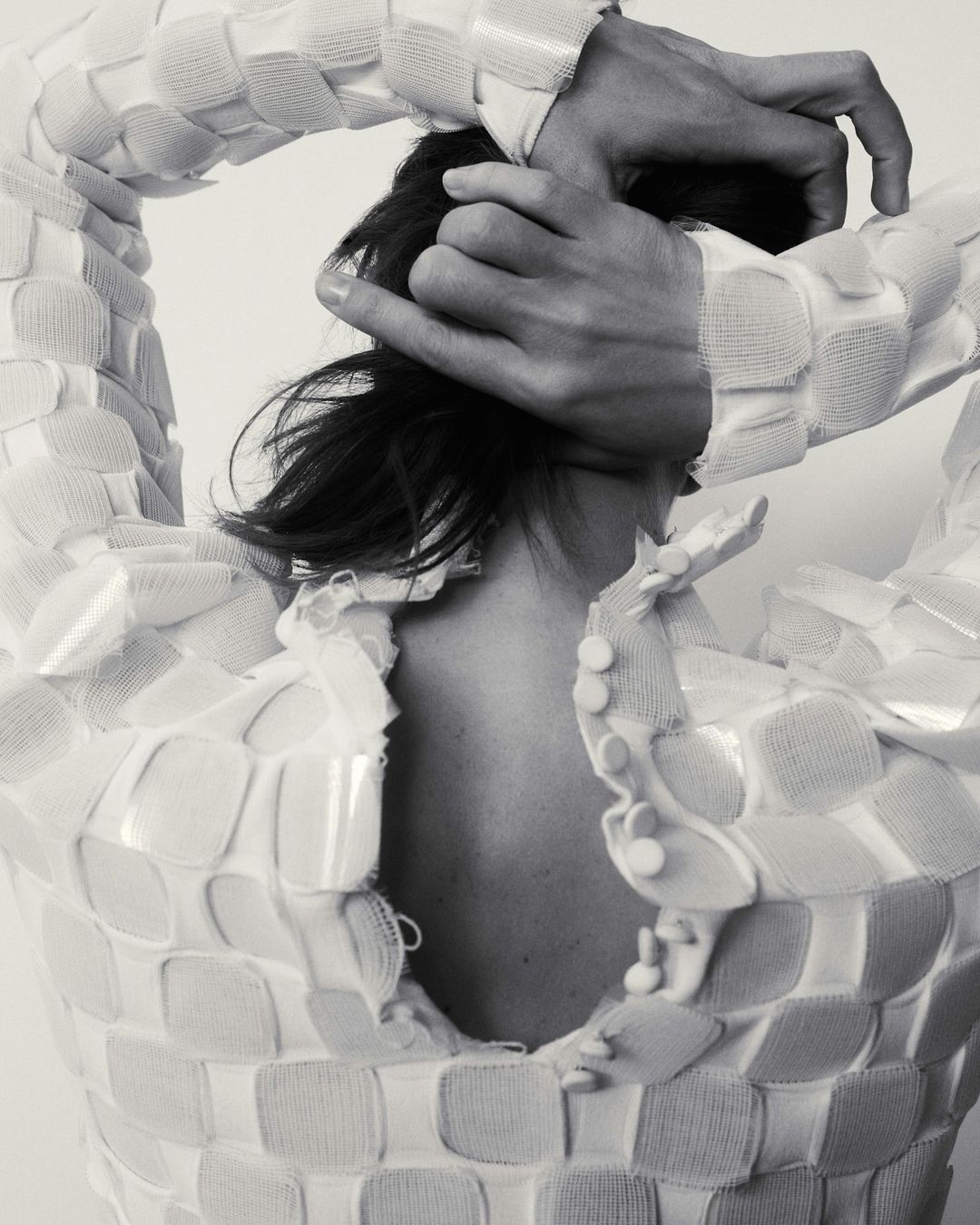 Monochrome studio shot of a person wearing a white woven garment, hands on head, back view, showing fashion and style details.