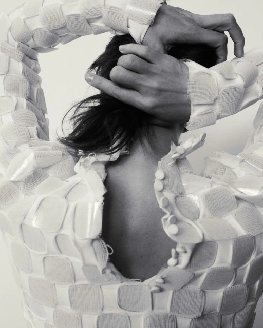 Monochrome studio shot of a person wearing a white woven garment, hands on head, back view, showing fashion and style details.