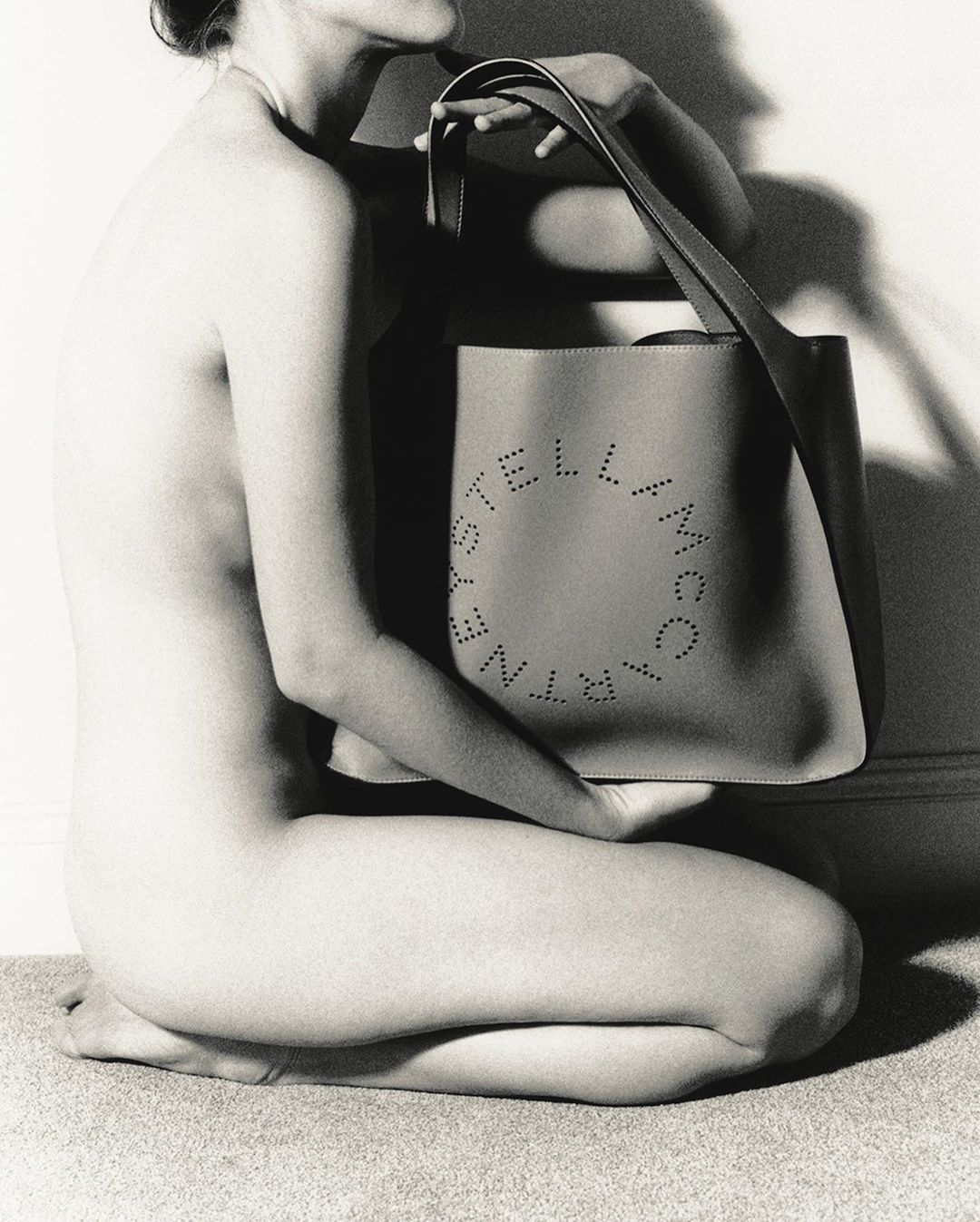Monochrome studio shot of a nude adult female kneeling while holding a Stella McCartney perforated tote bag against a white wall backdrop.