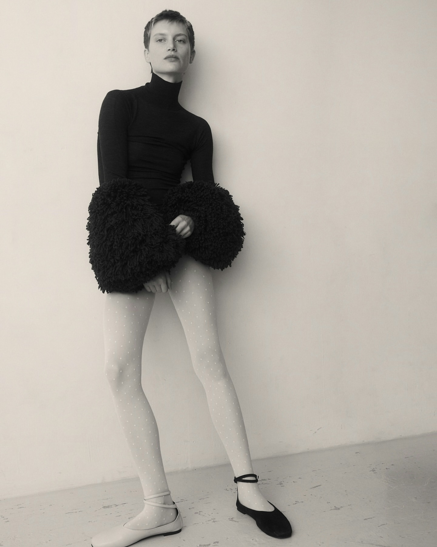 Monochrome fashion shot of a slim woman with short hair, wearing a black turtleneck, a textured skirt, and flats, against a neutral background.