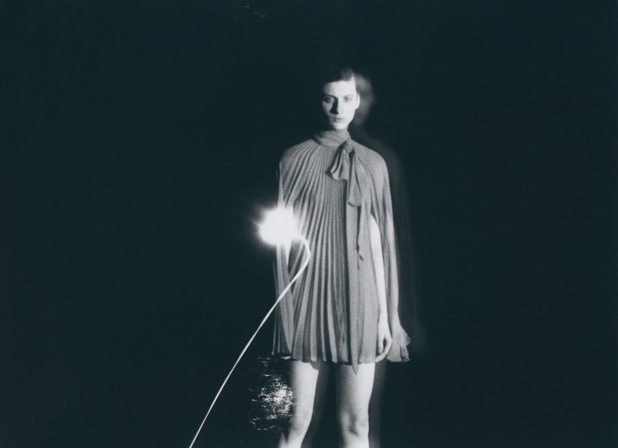 Monochromatic shot of a slender woman in a pleated dress, softly blurred, holding a glowing light source in a dark environment.