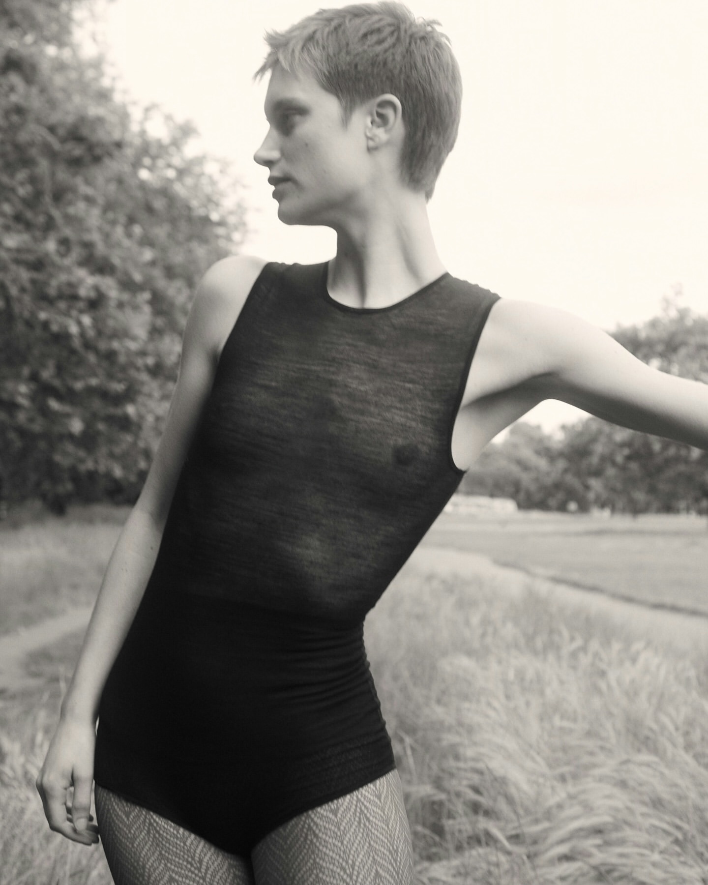 Woman poses outdoors in a black bodysuit. The landscape shows a field of tall grass and distant trees against a gray sky.
