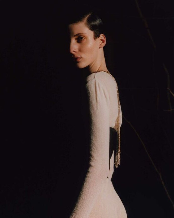 Portrait of a person with short hair wearing a light-colored dress and gold chain, standing against a dark background with subtle lighting.