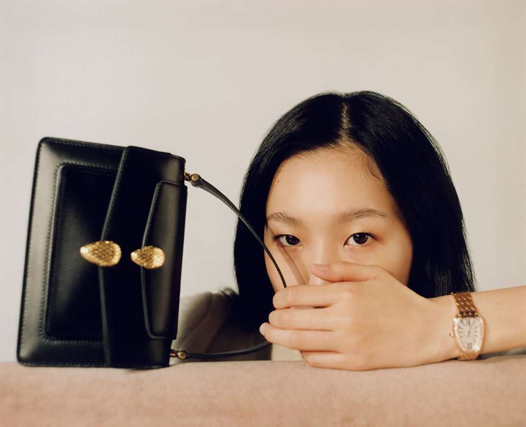 Asian woman poses indoors with a black leather handbag featuring golden hardware, complemented by her watch and a cream backdrop.