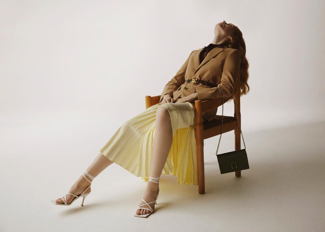 Woman in a blazer and pleated skirt reclines in a chair with a purse, showcasing fashion and style in a studio setting.