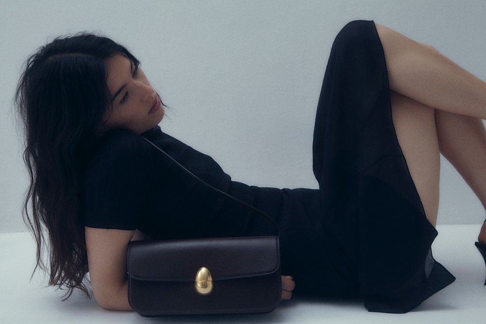 Woman in a black dress reclining on a white background with a dark leather shoulder bag in the foreground.