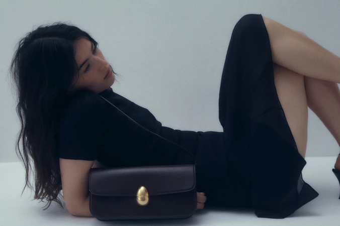 Woman in a black dress reclining on a white background with a dark leather shoulder bag in the foreground.