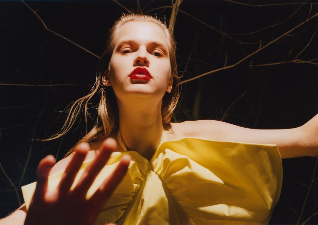 Blonde woman in yellow dress with red lipstick poses outdoors against a dark background with bare branches. Hand raised toward camera.