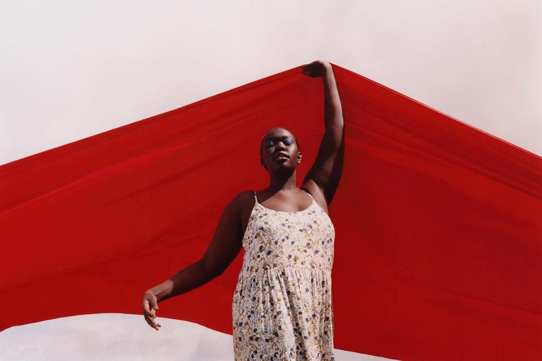 Black woman in a floral dress holding a vibrant red fabric against an overcast sky, captured by Daisy Walker.