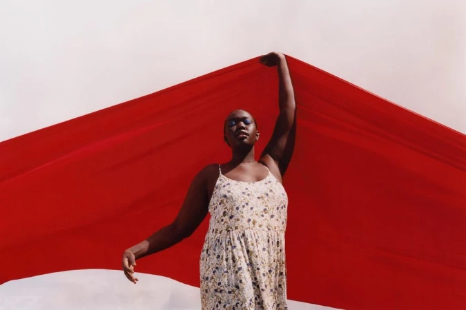 Black woman in a floral dress holding a vibrant red fabric against an overcast sky, captured by Daisy Walker.