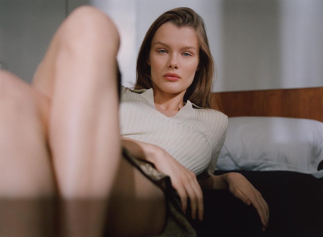 Portrait of a light-skinned woman with long brown hair lying on a bed wearing a cream-colored top in a softly lit indoor space.