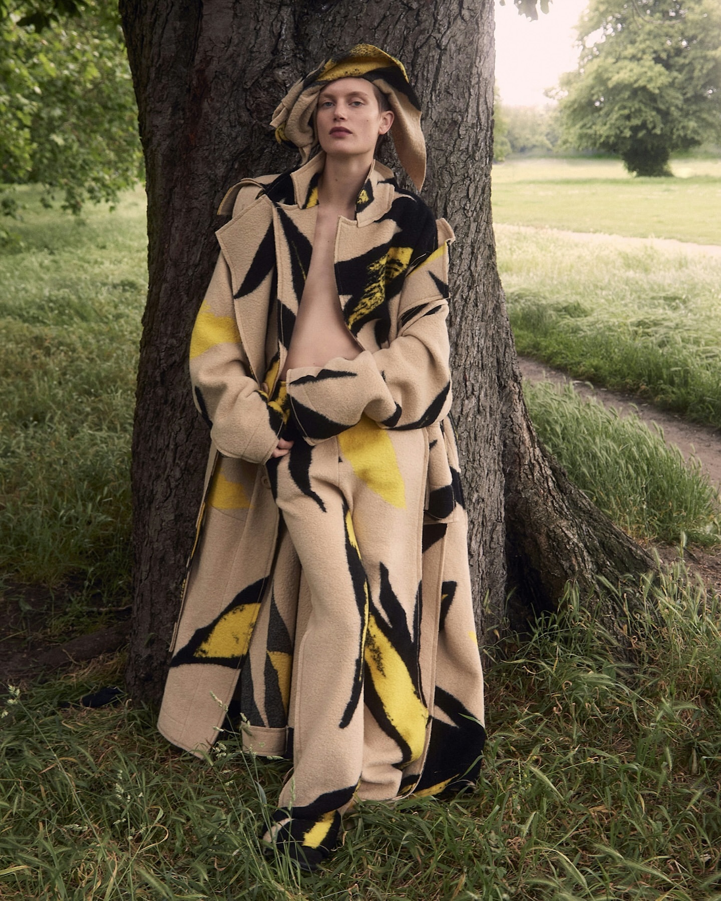 Adult female model stands next to a tree wearing a hat, coat, and pants with a black and yellow leaf pattern in a green grassy field.