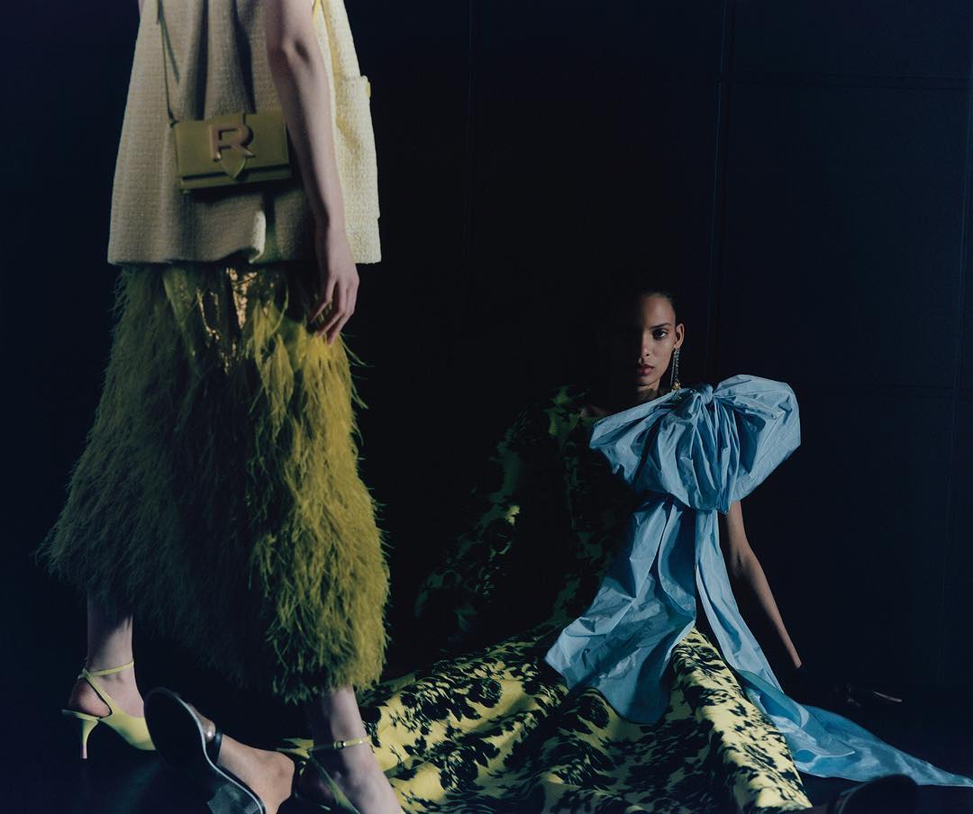Two fashion models pose against a dark background. One model wears a green feathered skirt and the other a blue bow dress.