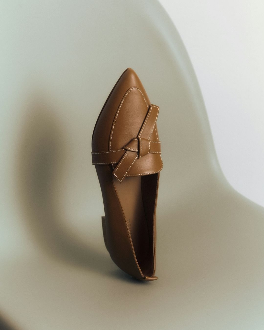 Studio shot of a tan leather loafer shoe with a pointed toe and a decorative knotted strap, set against a gradient white background.