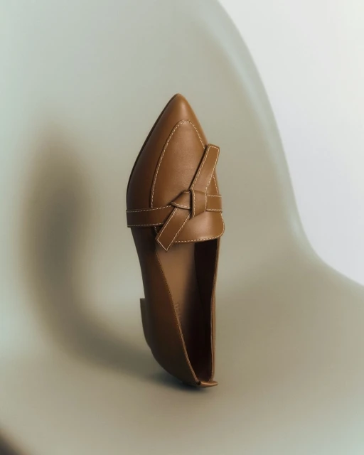 Studio shot of a tan leather loafer shoe with a pointed toe and a decorative knotted strap, set against a gradient white background.