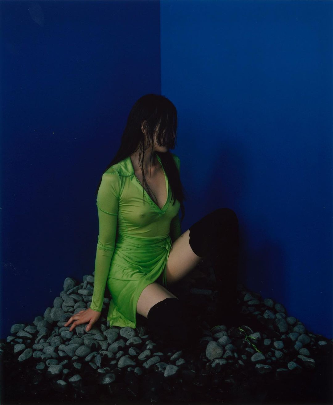 Woman in a vibrant green dress poses seated on a pile of rocks against a blue backdrop, highlighted by soft, diffused lighting.
