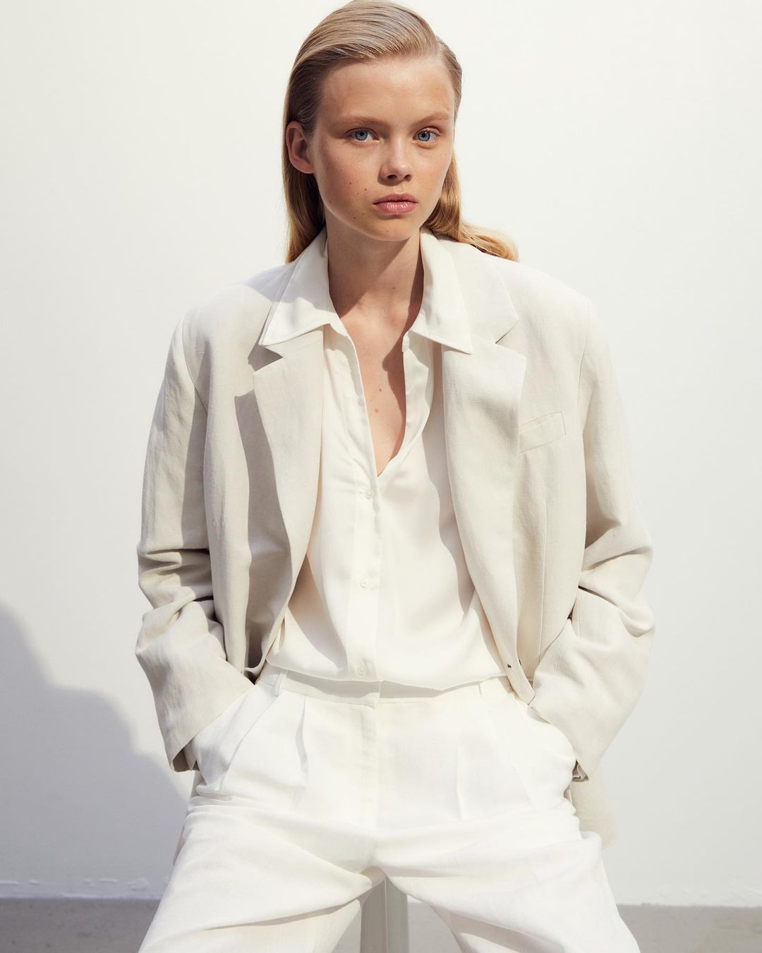 Fashion portrait of a blonde woman wearing an oversized linen blazer and trousers, set against a neutral background in a studio environment.