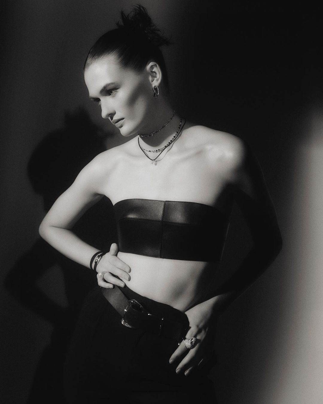 Monochrome portrait of a woman in a strapless top, posing with hands on her waist, wearing layered necklaces, earrings, and a belt.