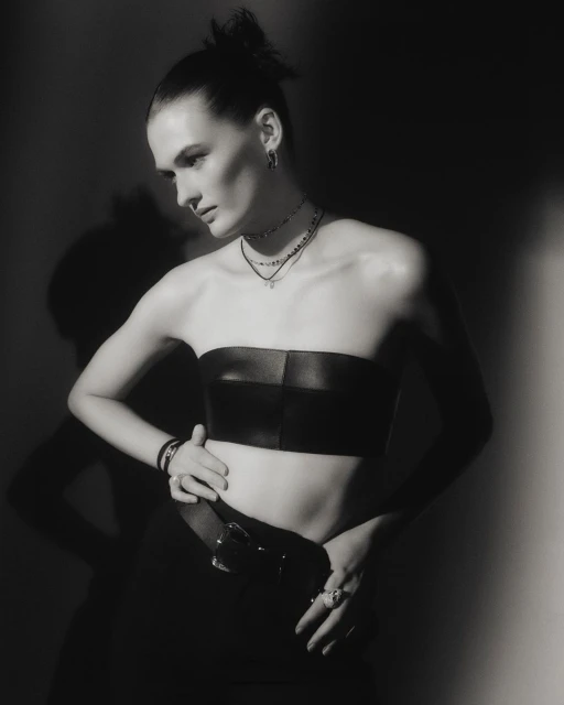 Monochrome portrait of a woman in a strapless top, posing with hands on her waist, wearing layered necklaces, earrings, and a belt.