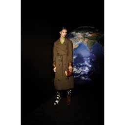 Model in a long brown belted trench coat holding a small handbag with a large globe backdrop by Daisy Walker