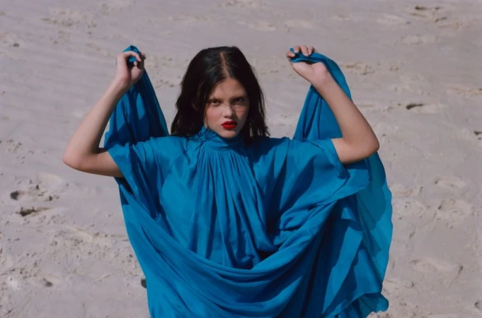 Woman in bright blue high neck dress holding fabric against sandy beach backdrop