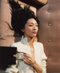 East Asian woman lying back on a tufted pink couch wearing a white knit top, holding a black handbag and wearing a watch.