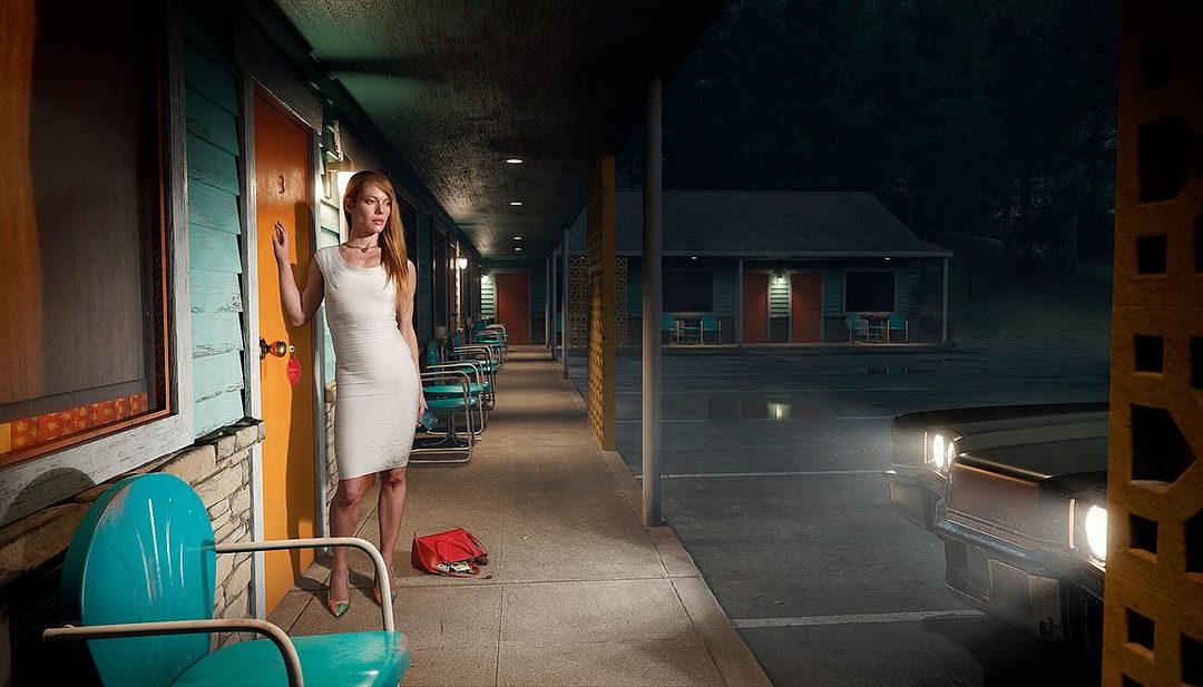 A stylish woman in a white dress stands in front of a retro motel at night, with vintage cars and chairs adding to the scene.