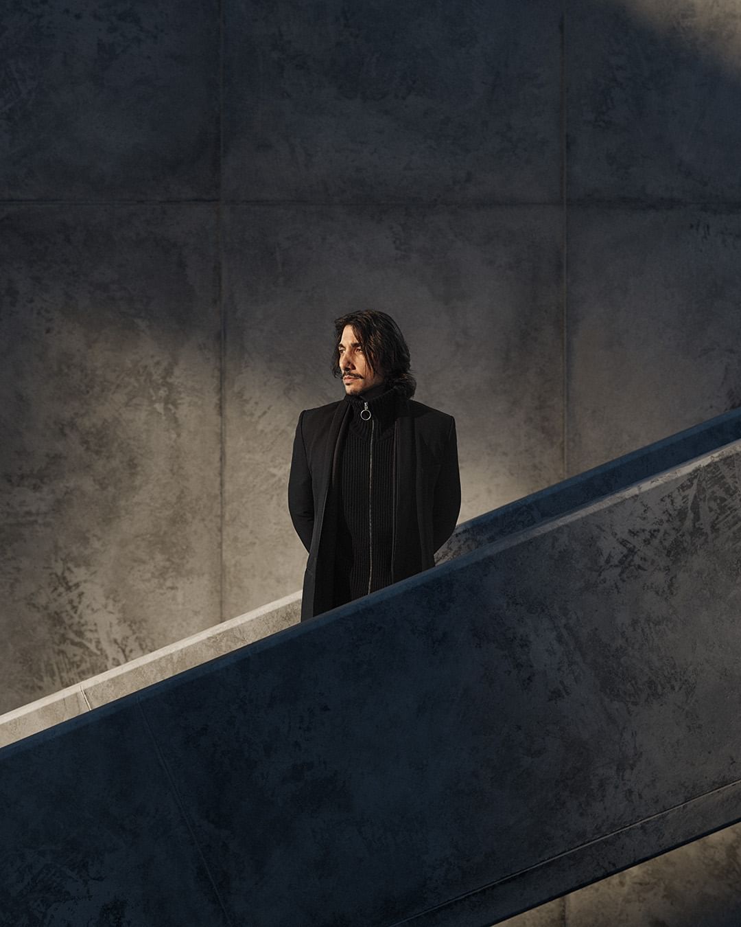 Portrait of an adult male with long dark hair, wearing a black coat, standing on a staircase against a concrete wall with shadows.