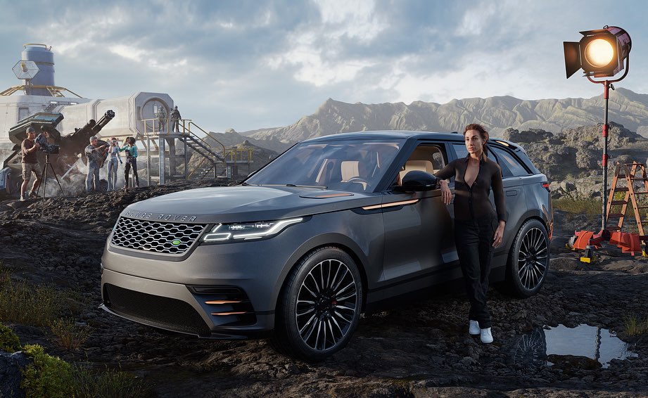 A gray Range Rover Velar, with an adult female model leaning on it, sits on a rocky landscape beside a studio lighting setup and crew.