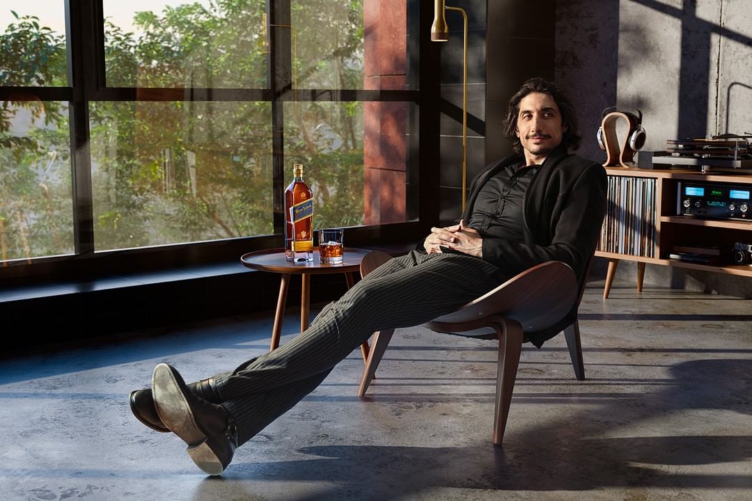 Adult male lounges in mid-century modern chair, wearing a black suit, by a window near liquor bottle and vintage audio equipment.