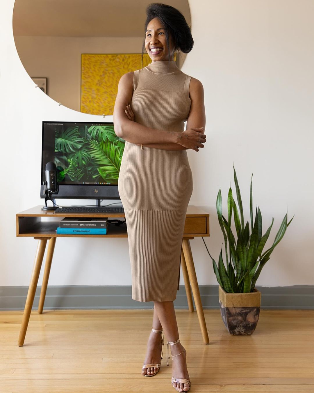 Elegant woman in a beige dress stands in a well-lit room with a desk, computer displaying green plants, and a decorative plant.