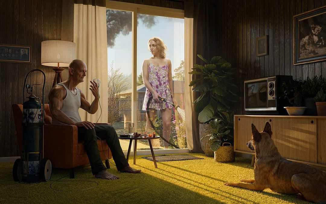 Indoor shot of a senior man connected to oxygen, seated in a chair, with a young woman watering the lawn outside, and a dog lying on the floor.