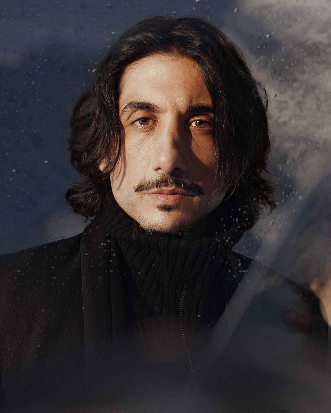 Portrait of a dark-haired adult male with a mustache wearing a black turtleneck sweater and jacket against a blurred window background.
