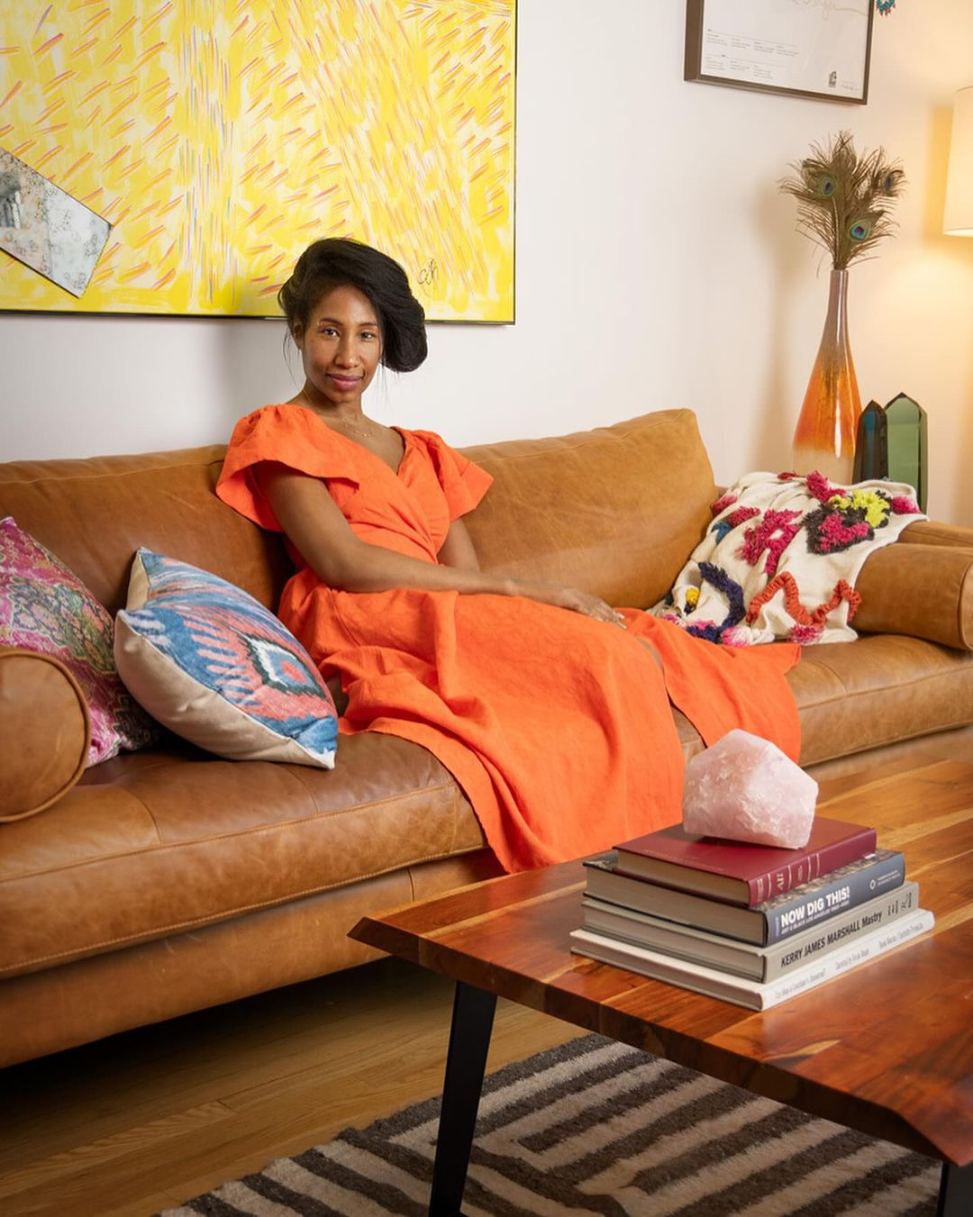 Woman in orange dress sits on a leather couch in a living room with colorful pillows, artwork, and a wooden coffee table.