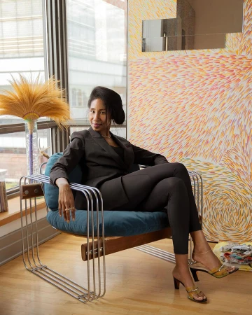Adult female in black business suit seated indoors in modern chair in room with art and natural light from a nearby window.