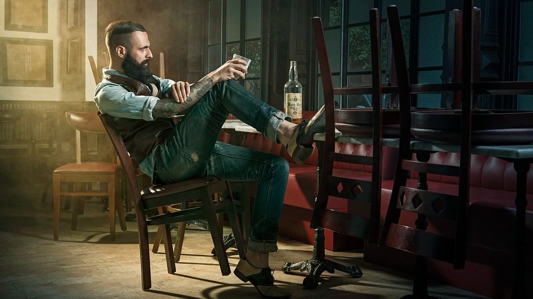 Bearded man in brown vest and blue jeans sits holding drink in bar interior with stacked chairs, bottle, warm and cool color grading.