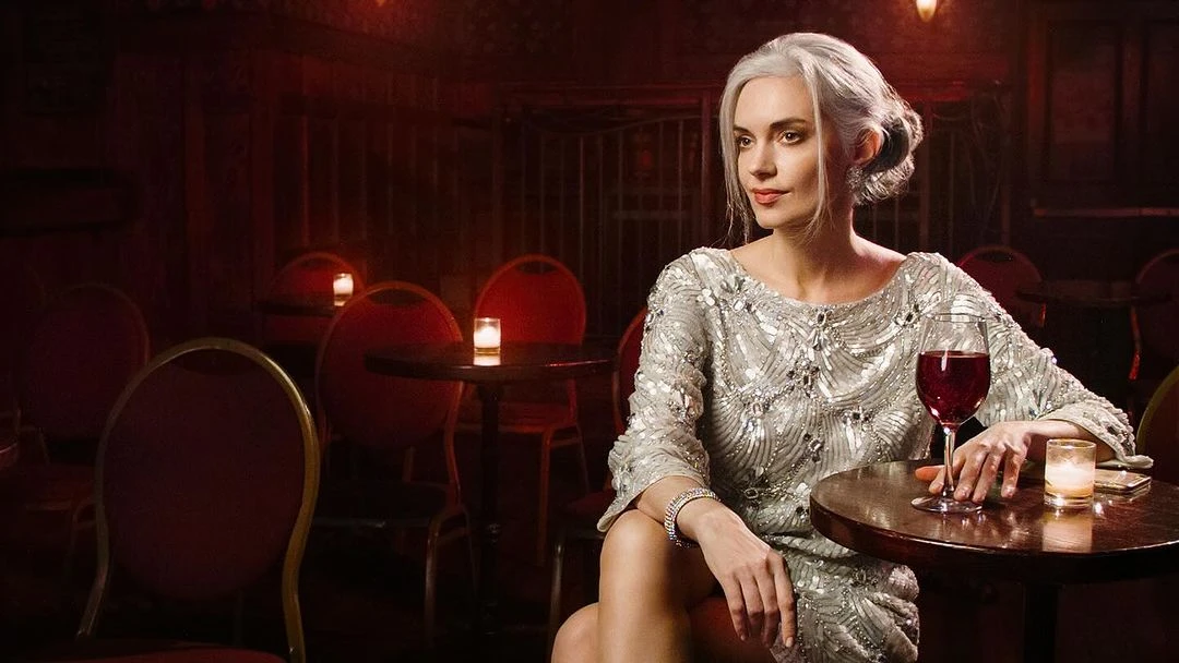 Elegant woman in silver dress sits at table in dimly lit restaurant with wine glass, creating a vintage, glamorous aesthetic.