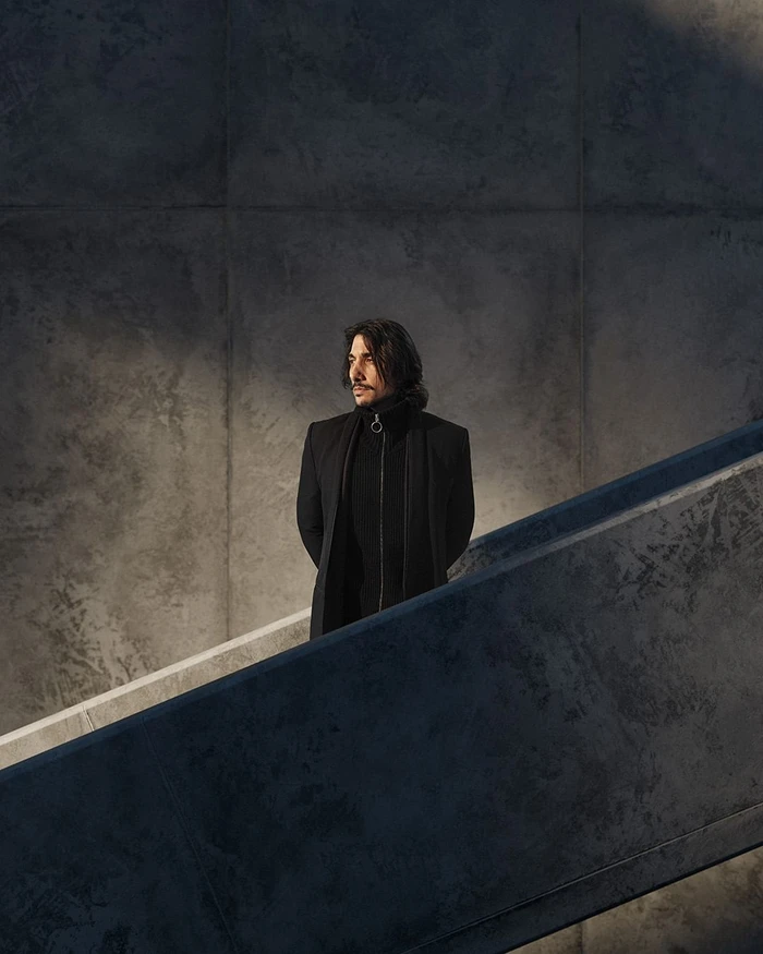 Man with long dark hair wearing a black coat standing on a concrete staircase in New York by Dale May