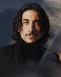 Portrait of a man with long dark hair, mustache, and goatee wearing a black turtleneck and scarf, photographed by Dale May.