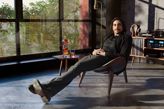 Man seated comfortably in a modern wooden lounge chair holding a glass of amber whiskey with a bottle on a side table.