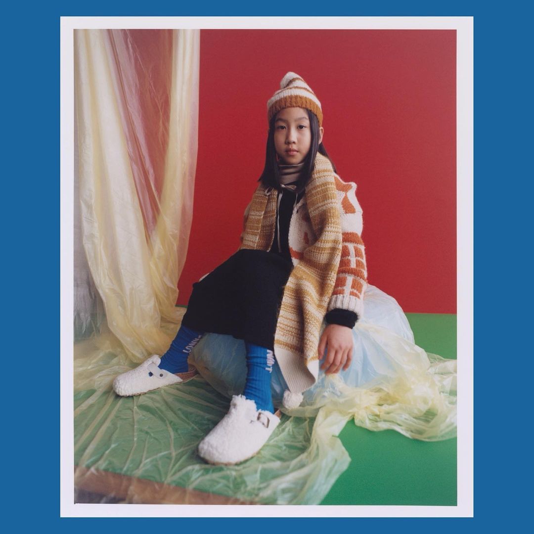 Young child poses in a warm knitted hat and cardigan, sitting on a draped surface against a contrasting red and green backdrop in a studio.