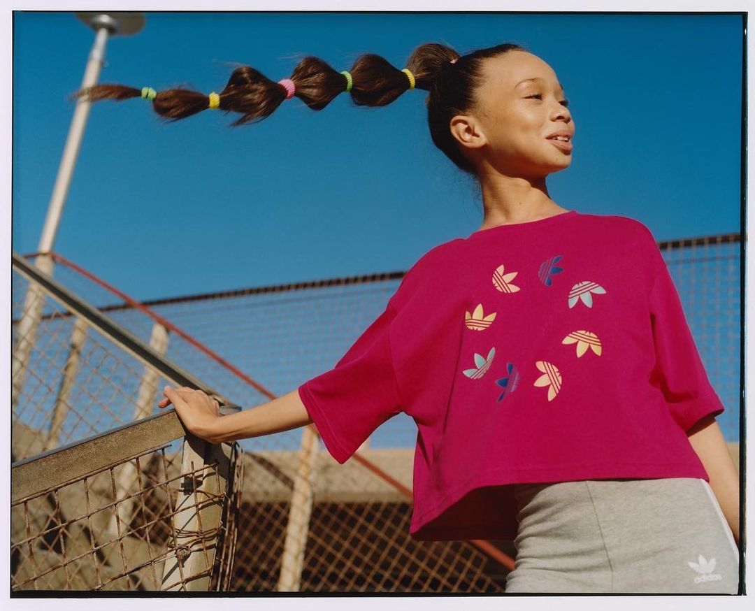 Teen girl in pink Adidas shirt with ponytail and colorful hair ties poses outdoors against blue sky and metal barrier in natural light.