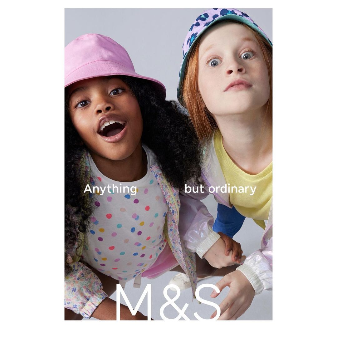 Two children, an African-American girl with a pink hat and a red-haired girl with a patterned hat, pose in a bright indoor studio.